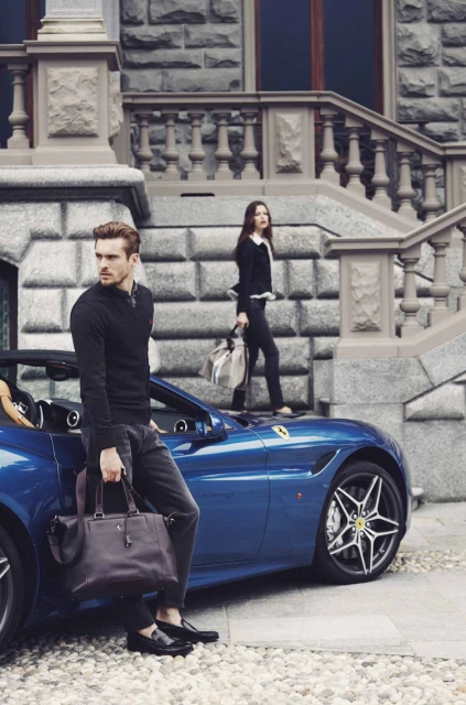 Well-dressed man with leather bag poses near a blue convertible car with a woman on a stone staircase in the background.