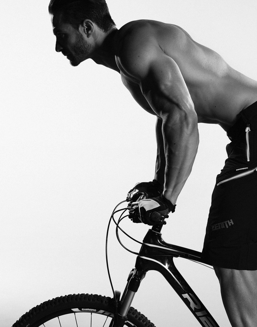 A muscular man leans forward while holding the handlebars of a mountain bike against a bright background in a black-and-white studio shot.