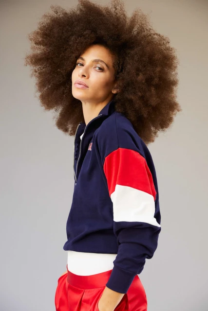 A woman with an afro hairstyle poses indoors wearing a color-blocked zip-up jacket and shiny red pants against a neutral backdrop.