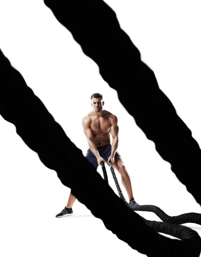 Muscular man works out with battle ropes against a white background in a fitness studio, illuminated by bright lighting.