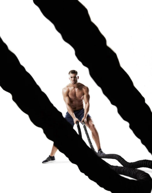 Muscular man works out with battle ropes against a white background in a fitness studio, illuminated by bright lighting.