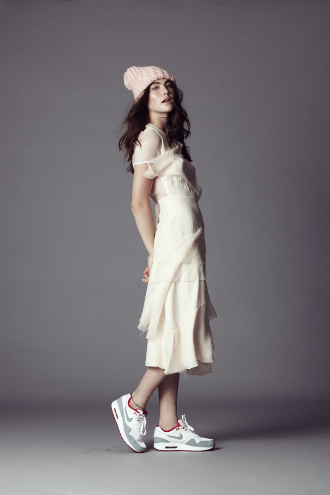 A young woman wearing a beanie and a long dress stands in a photo studio wearing running shoes, striking a fashion pose, and looking sideways.