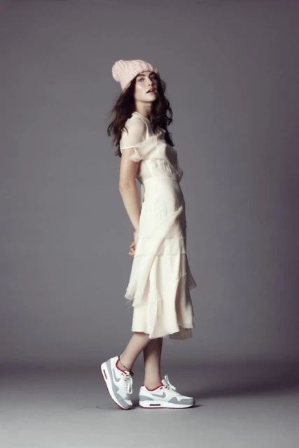 A young woman wearing a beanie and a long dress stands in a photo studio wearing running shoes, striking a fashion pose, and looking sideways.