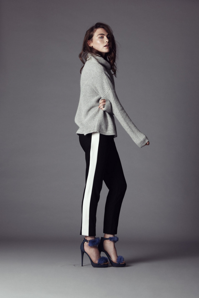 Fashion shot of a woman in a grey sweater and black pants with a white stripe posing in a studio setting against a grey backdrop.