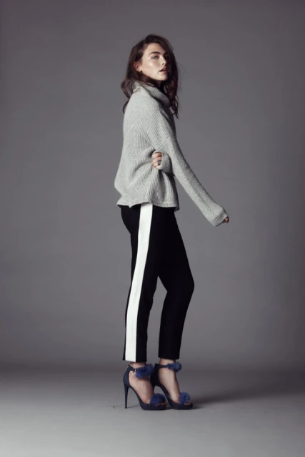 Fashion shot of a woman in a grey sweater and black pants with a white stripe posing in a studio setting against a grey backdrop.