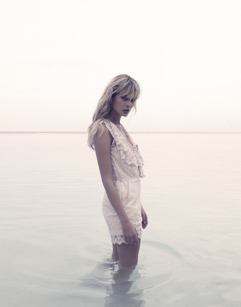 A solo woman stands in shallow water on a bright day, wearing a lacy off-white romper, with her blonde hair down and tousled.
