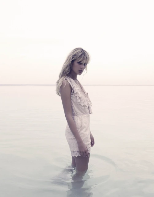 A solo woman stands in shallow water on a bright day, wearing a lacy off-white romper, with her blonde hair down and tousled.