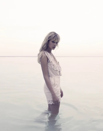 A solo woman stands in shallow water on a bright day, wearing a lacy off-white romper, with her blonde hair down and tousled.