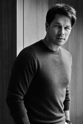 Portrait of a man wearing a dark crewneck sweater standing indoors by a paneled wooden wall.