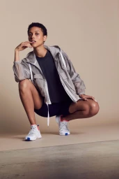 Model squatting in a grey hooded windbreaker and dark shorts against a neutral beige background.