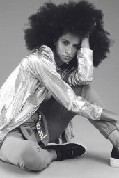 Black woman with voluminous afro hair wearing metallic jacket and sneakers seated against light background