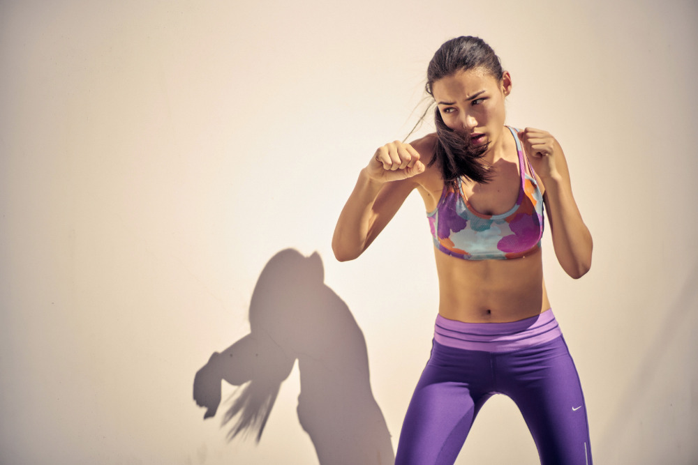 Athletic woman in sportswear strikes a boxing pose against a light wall, showcasing fitness and determination in a sunlit indoor setting.