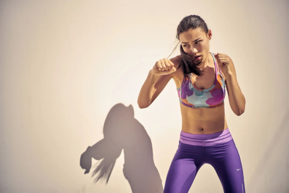 Athletic woman in sportswear strikes a boxing pose against a light wall, showcasing fitness and determination in a sunlit indoor setting.