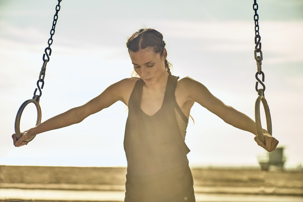 A woman in athletic wear hangs from gymnastics rings in an outdoor fitness setting with a bright sky and a blurred background.