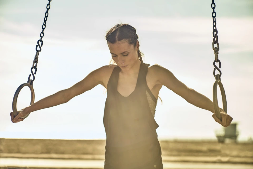A woman in athletic wear hangs from gymnastics rings in an outdoor fitness setting with a bright sky and a blurred background.