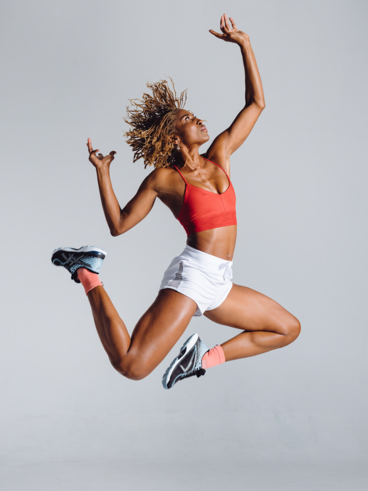 Athletic woman with blonde afro in red top and white shorts leaping mid-air in a studio setting, hands raised above her head.