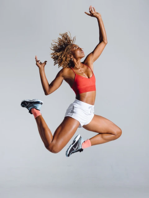 Athletic woman with blonde afro in red top and white shorts leaping mid-air in a studio setting, hands raised above her head.