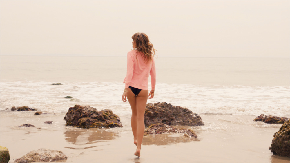 Woman walking on a sandy beach wearing a pink top and dark bottoms with waves and rocks in the background