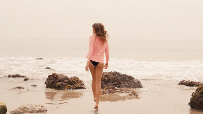 Woman walking on a sandy beach wearing a pink top and dark bottoms with waves and rocks in the background