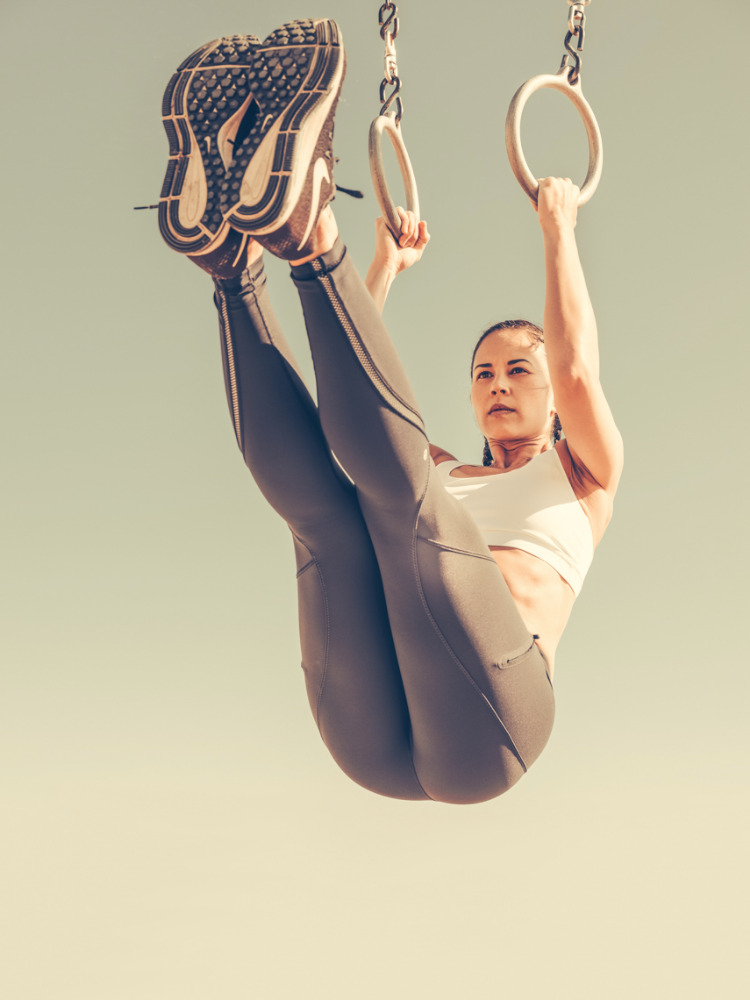 Athletic woman performing exercise on gymnastic rings in outdoor setting, wearing a white sports bra and grey leggings against a blue sky.