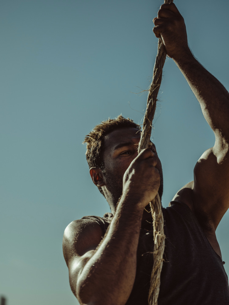 African-American man with dark tank top climbing thick rope under bright sunlight against clear blue sky showing arm muscle definition.