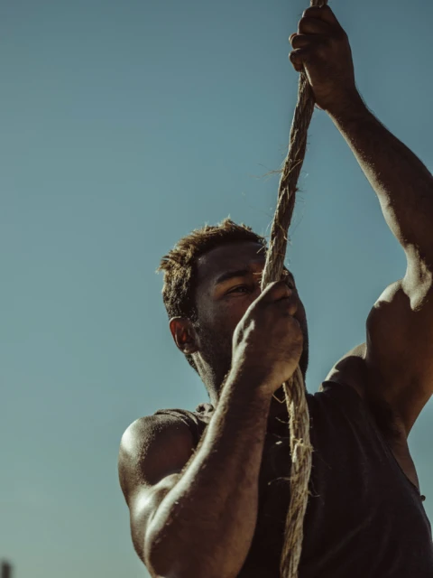 African-American man with dark tank top climbing thick rope under bright sunlight against clear blue sky showing arm muscle definition.