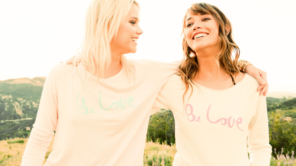 Two smiling young women embrace in a sun-drenched field, wearing matching 'Be Love' sweatshirts, with soft focus hills in background.