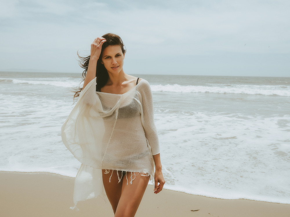 A woman poses on a sandy beach near the ocean wearing a light, sheer cover-up, enjoying a breezy day with gentle waves in the background.
