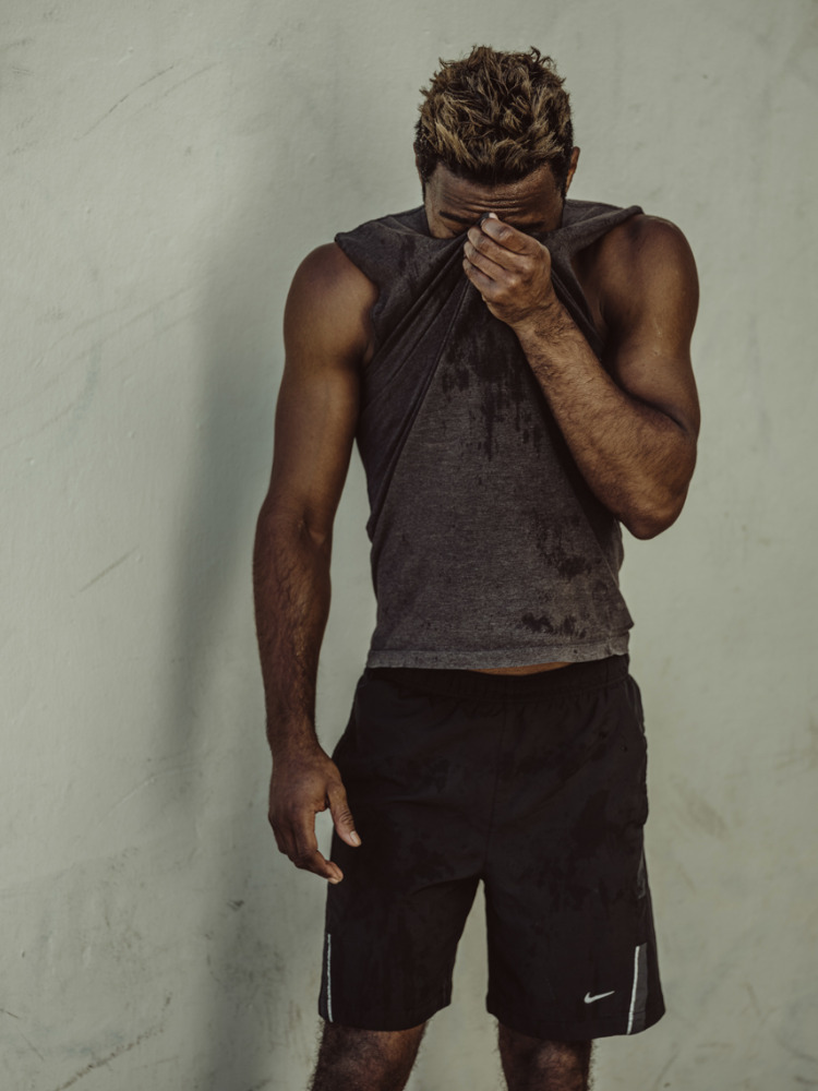 Athletic, sweaty adult male in black shorts wipes his face with his gray t-shirt in front of a textured gray wall with side lighting.