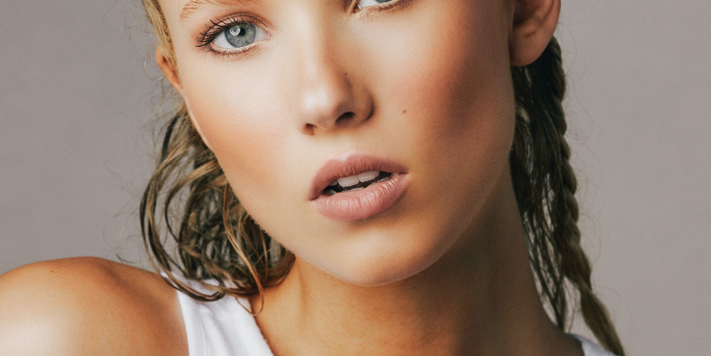 Close-up portrait of a woman with light skin, blonde hair in braids, light blue eyes, and light pink lips against a neutral background.