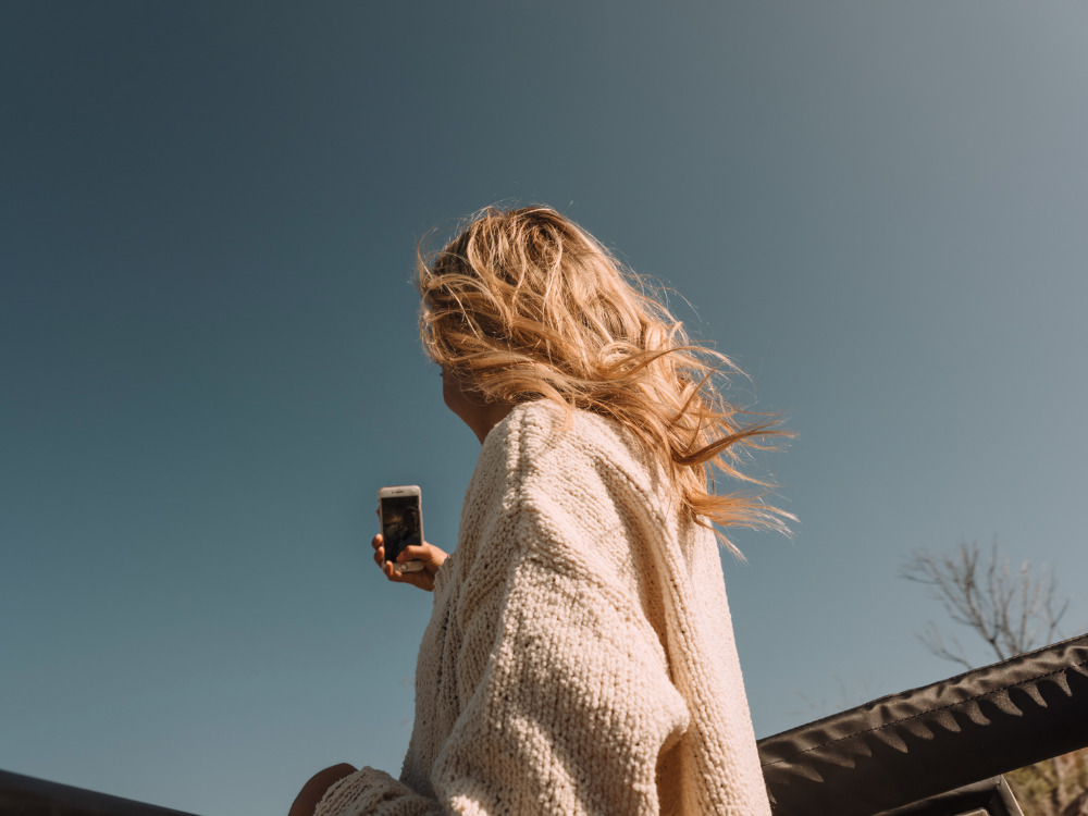 Blonde woman wearing a cream knitted sweater holding a smartphone against a clear blue sky by Paul Schefz.