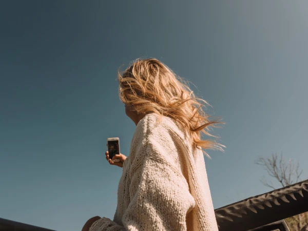 Blonde woman wearing a cream knitted sweater holding a smartphone against a clear blue sky by Paul Schefz.