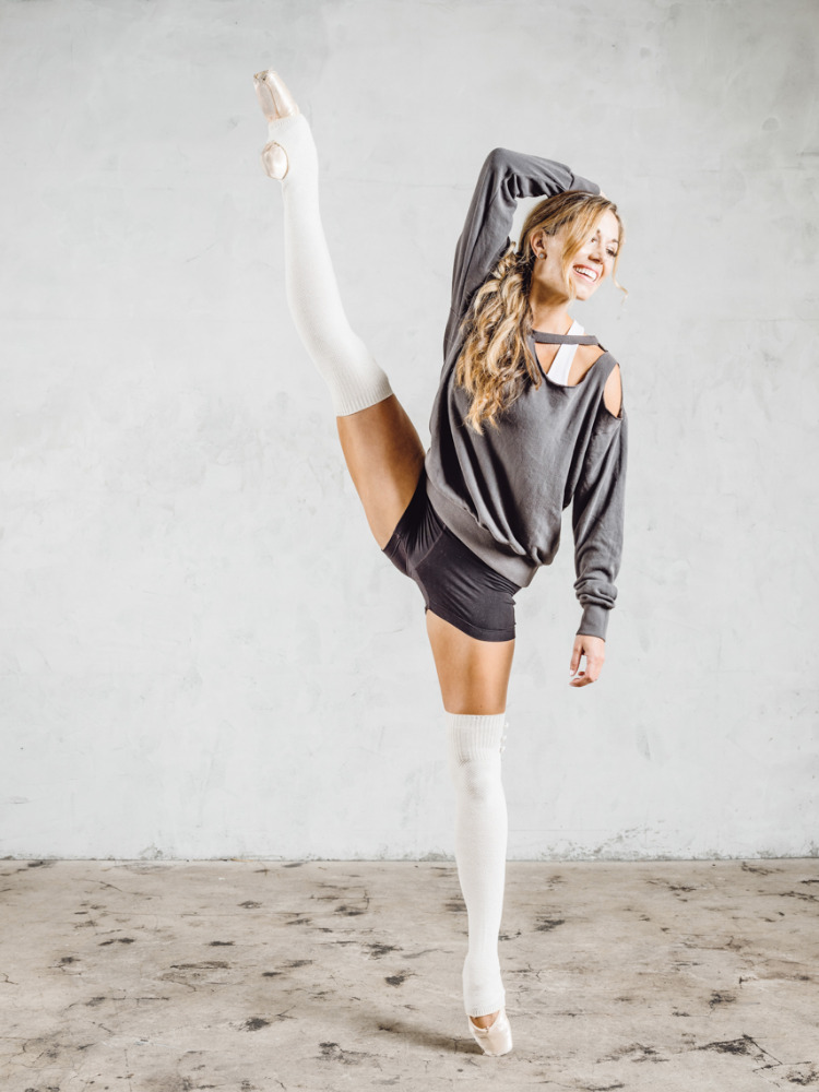 A woman in dance wear balances while holding a leg up during a dance pose inside a photo studio with a neutral backdrop.