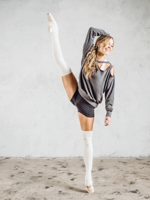 A woman in dance wear balances while holding a leg up during a dance pose inside a photo studio with a neutral backdrop.