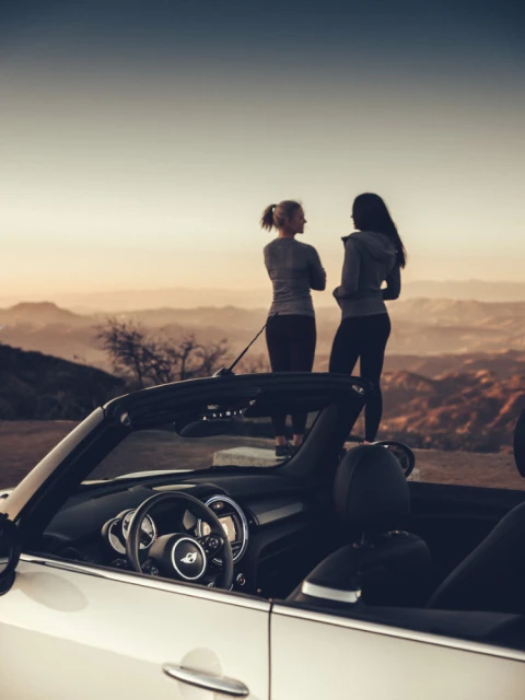Two women standing near a white convertible car on a scenic overlook at sunset with rolling hills in the background, captured at eye-level.