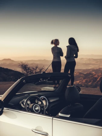 Two women standing near a white convertible car on a scenic overlook at sunset with rolling hills in the background, captured at eye-level.