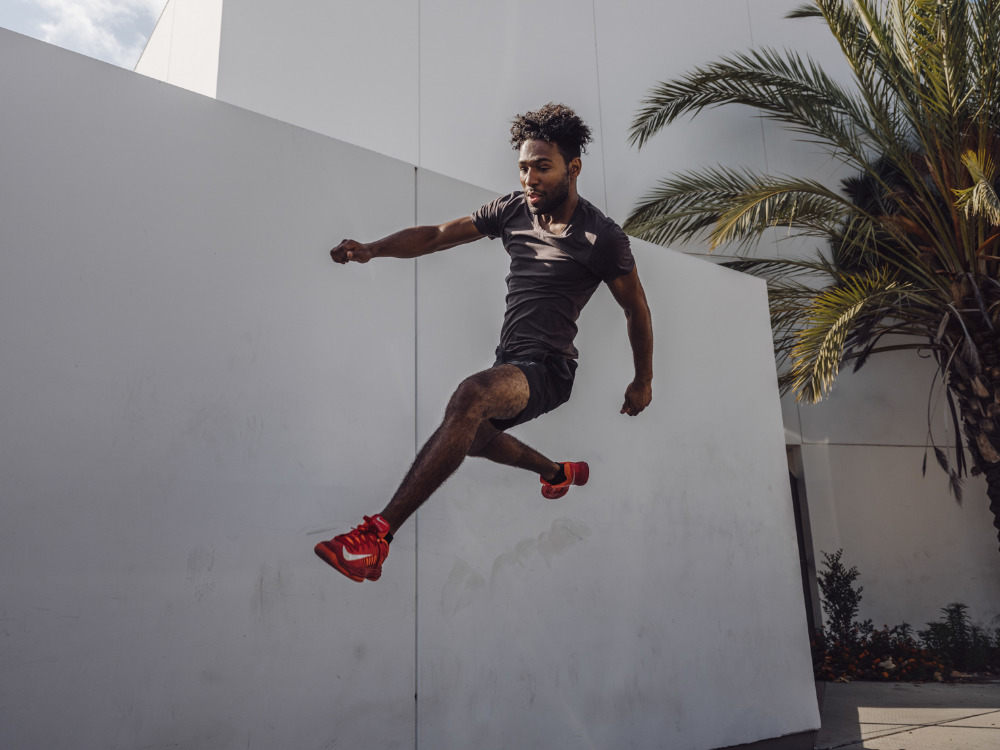 Athletic Black man in black activewear leaps high against a white building and palm tree on a sunny day, showcasing fitness.