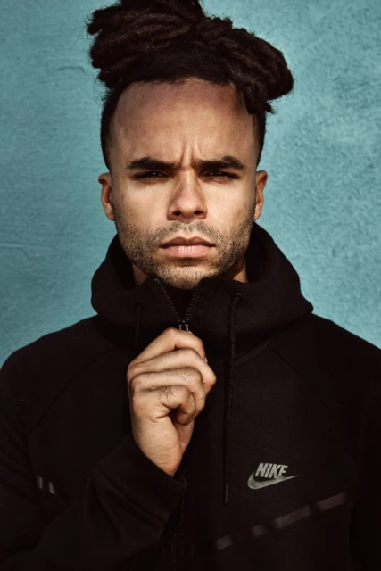 Portrait of an adult male with dreadlocks wearing a black Nike hooded jacket against a textured blue wall background indoors.