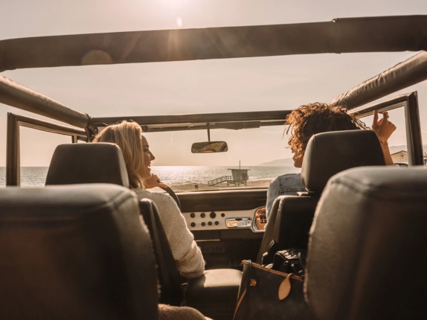 Two women enjoy a sunny beach drive in an open-top jeep with ocean views and a distant lifeguard tower, capturing a relaxed, summery road trip.