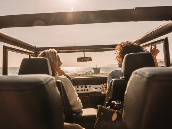 Two women enjoy a sunny beach drive in an open-top jeep with ocean views and a distant lifeguard tower, capturing a relaxed, summery road trip.