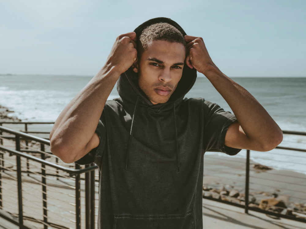 Man in a dark hooded t-shirt standing near a walkway with the beach and the ocean in the background on a sunny day.