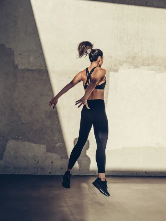 Woman wearing black athletic wear jumping in mid-air against a textured concrete wall with dramatic diagonal lighting.
