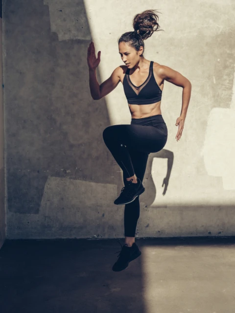 Athletic woman in black sports bra and leggings jumps with knee raised in a fitness workout indoors against a textured wall.