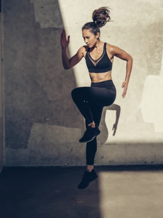 Female athlete captured mid-stride running in black activewear against a textured concrete wall with stark shadows.