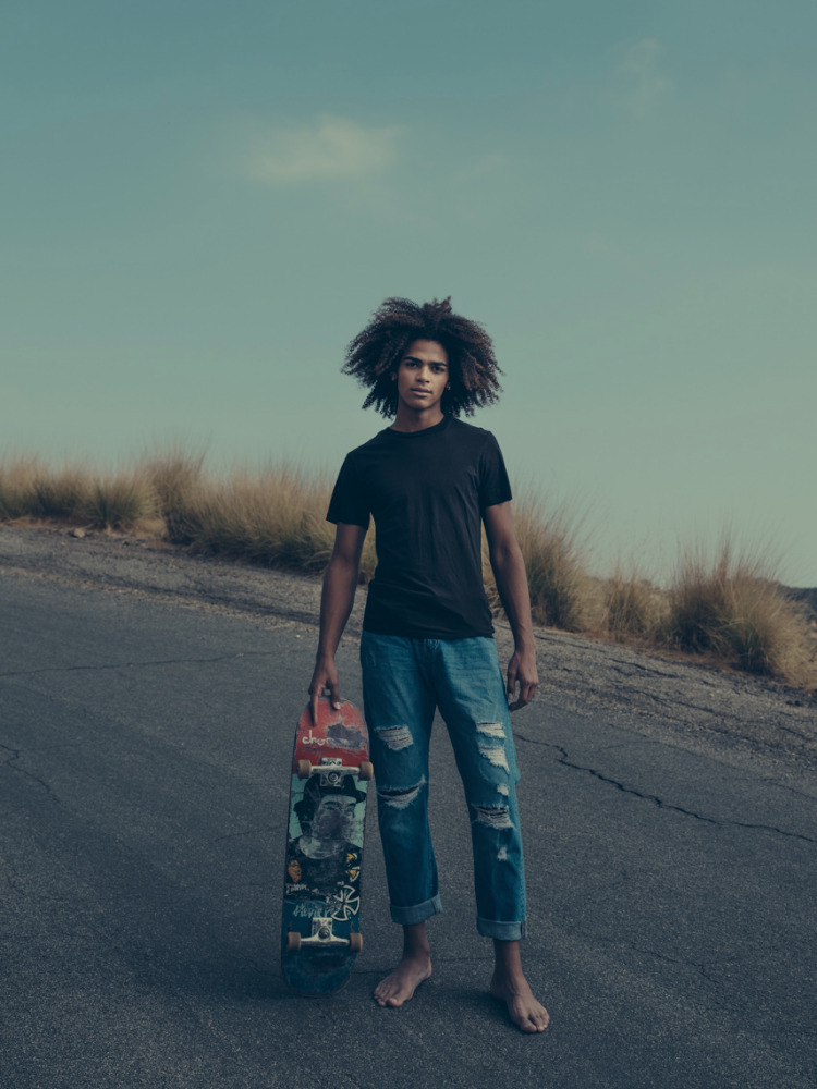 Young man with afro stands barefoot with a skateboard on an empty road with dry grassy hills and a blue sky on the horizon.