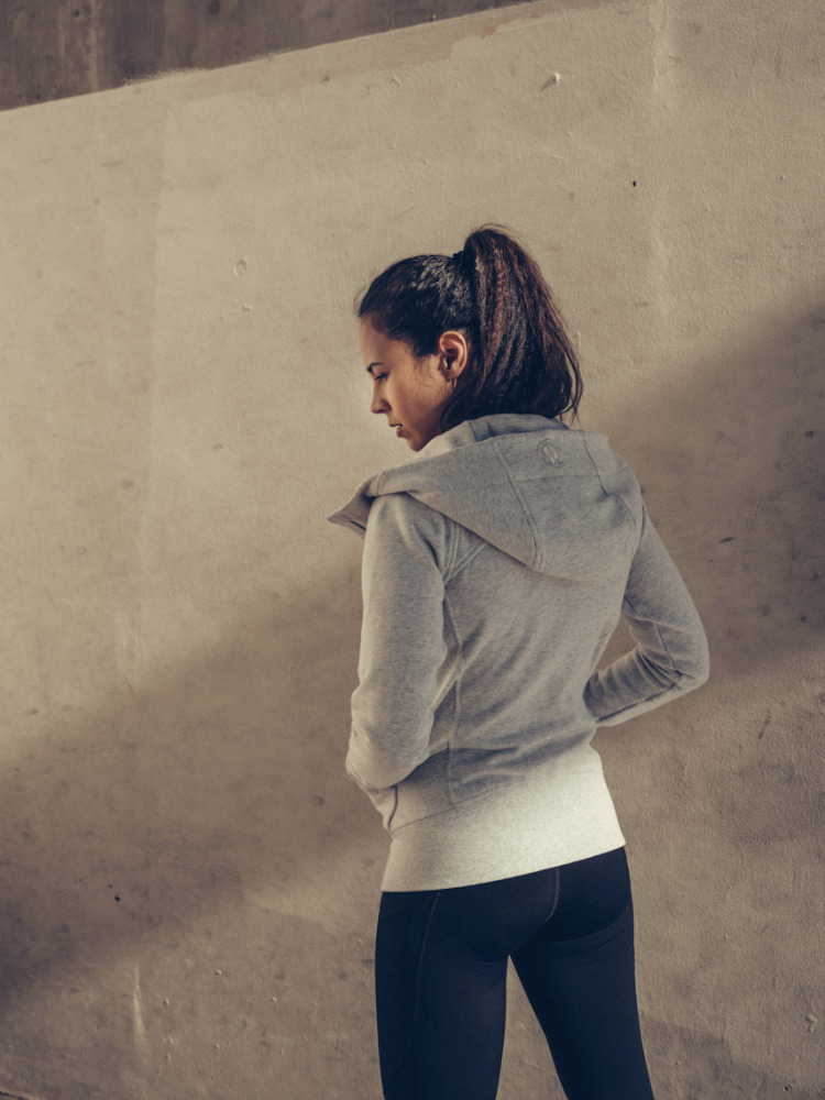 Back view of woman in grey athletic hoodie and leggings standing in front of a concrete wall, posing in an indoor environment.