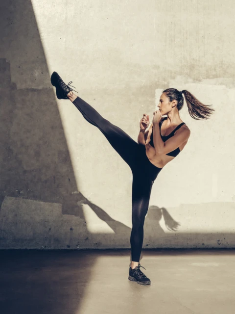 Athletic woman in black activewear powerfully performs a high kick in a brightly lit, minimalist indoor studio setting.