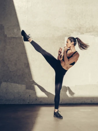 Athletic woman in black activewear powerfully performs a high kick in a brightly lit, minimalist indoor studio setting.