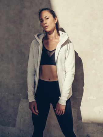 Woman wearing a grey zip hoodie and black athletic wear standing against a textured concrete wall.