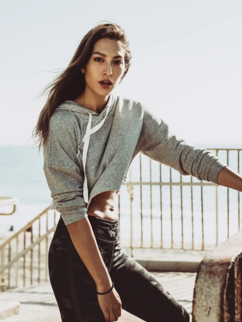 Athletic woman poses in gray hoodie and camouflage leggings by a beach railing on a sunny day, exuding health and fitness.